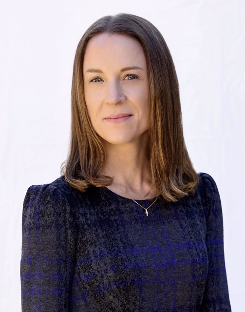 Portrait of a woman with straight, shoulder-length brown hair, wearing a textured dark dress with subtle blue tones and a delicate gold necklace. She stands against a softly lit white background, looking calmly at the camera with a slight smile.