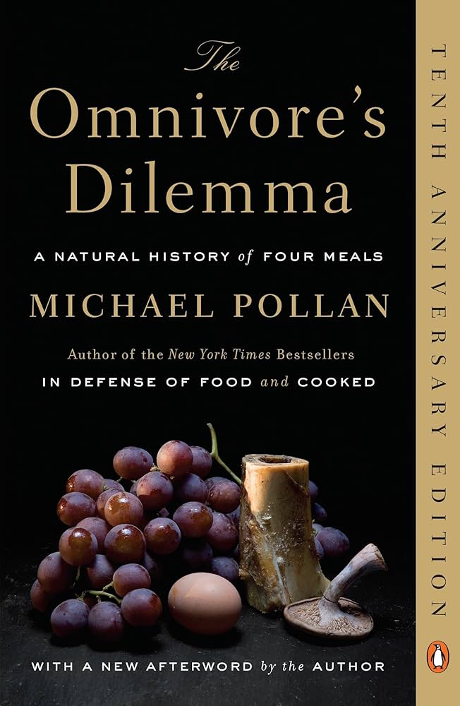 Book cover for The Omnivore's Dilemma by Michael Pollan, Tenth Anniversary Edition. A still-life arrangement of grapes, an egg, a bone, and a mushroom sits against a black background.