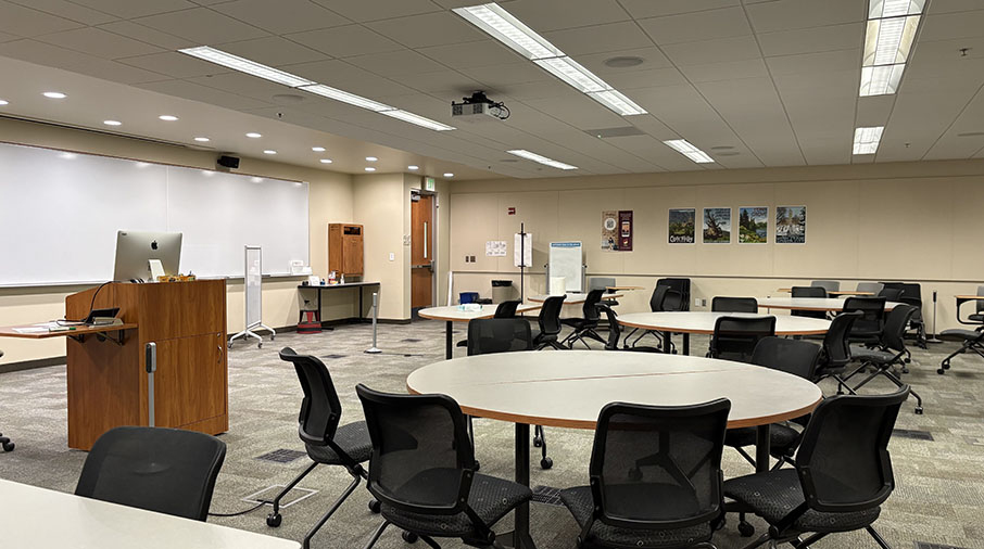 Classroom 122 - contains round tables and chairs, front of classroom has a whiteboard and projector capable.