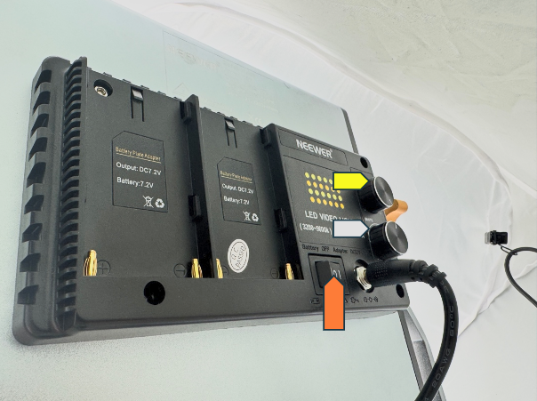 The rear view of a Neewer LED video light, showing two battery mounts and control knobs. A yellow arrow points to the brightness/color adjustment knobs, and an orange arrow points to the power switch near the DC input cable.